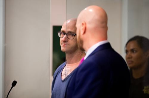 A bald man in glasses and blue jail clothes stands in an booth in a courtroom