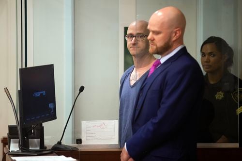 A bald man in glasses and blue jail clothes stands in an booth in a courtroom