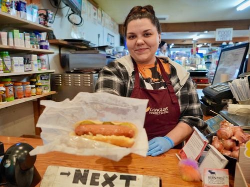a woman behind a deli counter holds out a hot dog wrapped in paper