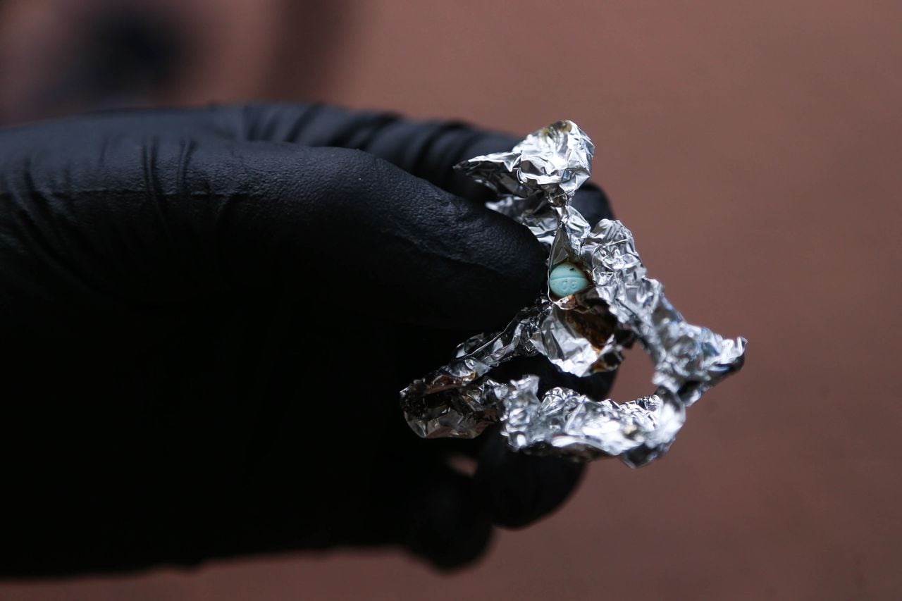 A hand clad in a black glove holds tin foil containing a blue pill.