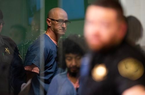 A bald man in glasses and blue jail clothes is escorted out of the back of a courtroom